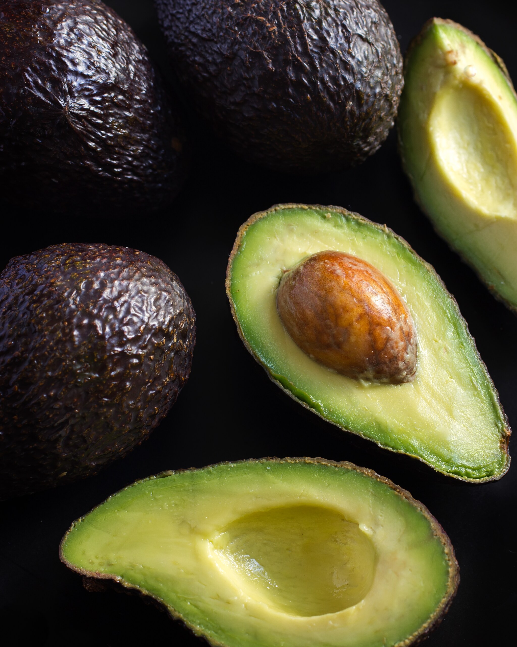 Whole avocados and two halves, one of them still holding its stone, on a black surface seen from above.