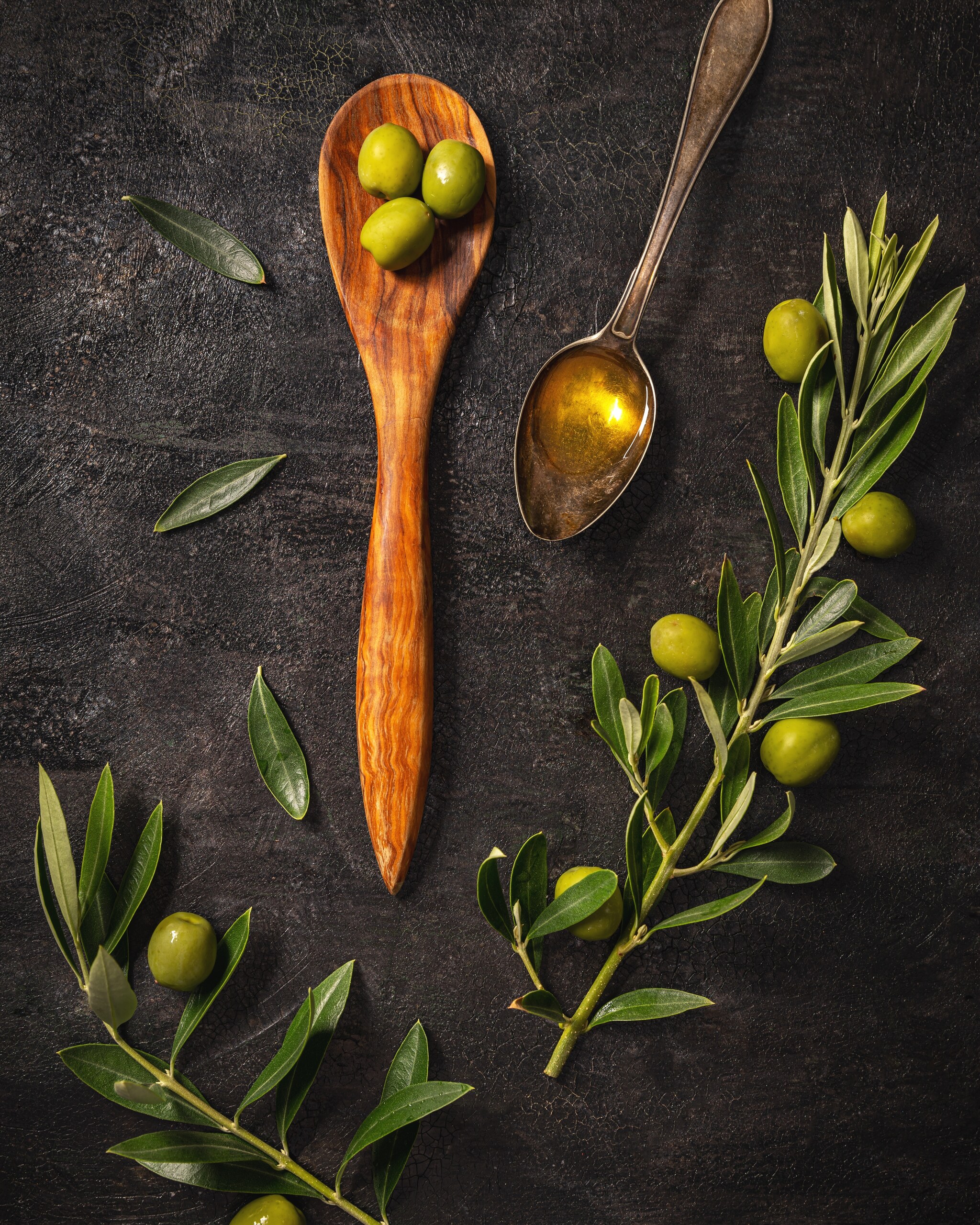Olive branches, loose green olives, a wooden spoon holding olives and a metal spoon holding olive oil, arranged on a dark surface and seen from directly above.