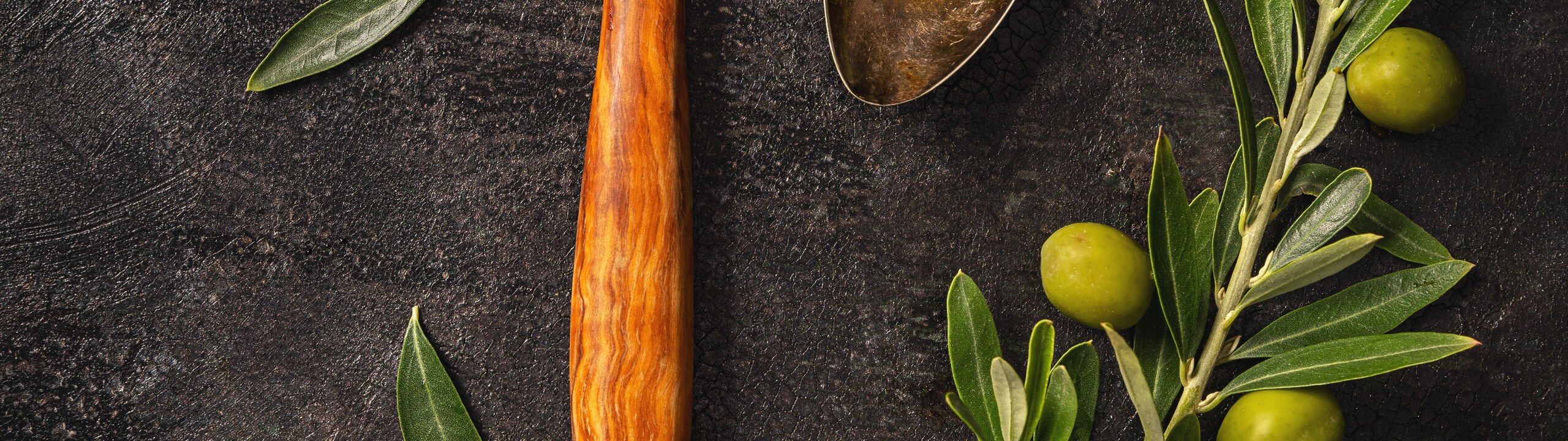 Olive branches, loose green olives, a wooden spoon holding olives and a metal spoon holding olive oil, arranged on a dark surface and seen from directly above.