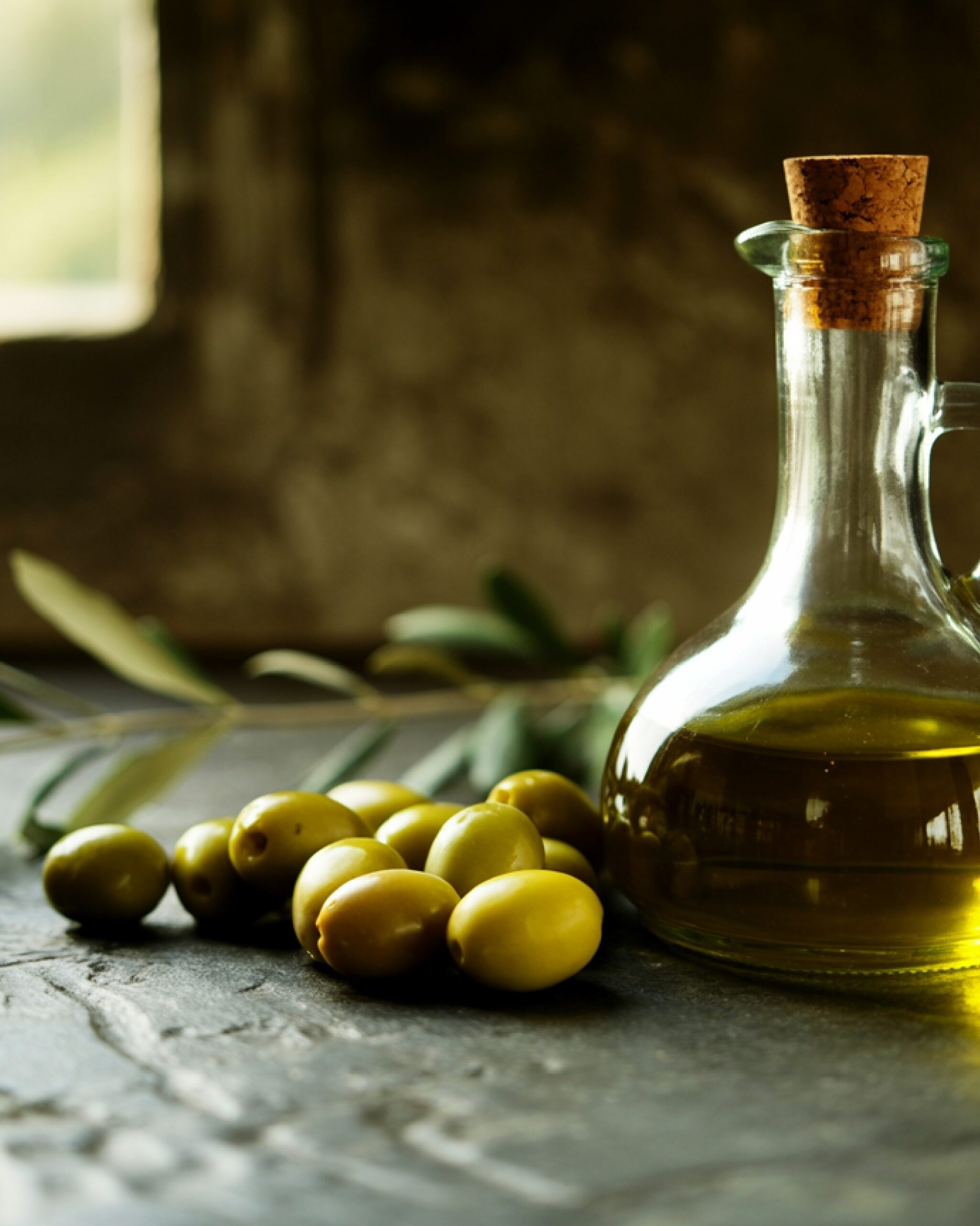 A glass cruet of olive oil beside a small heap of green olives and an olive branch, on a stone surface in soft window light.