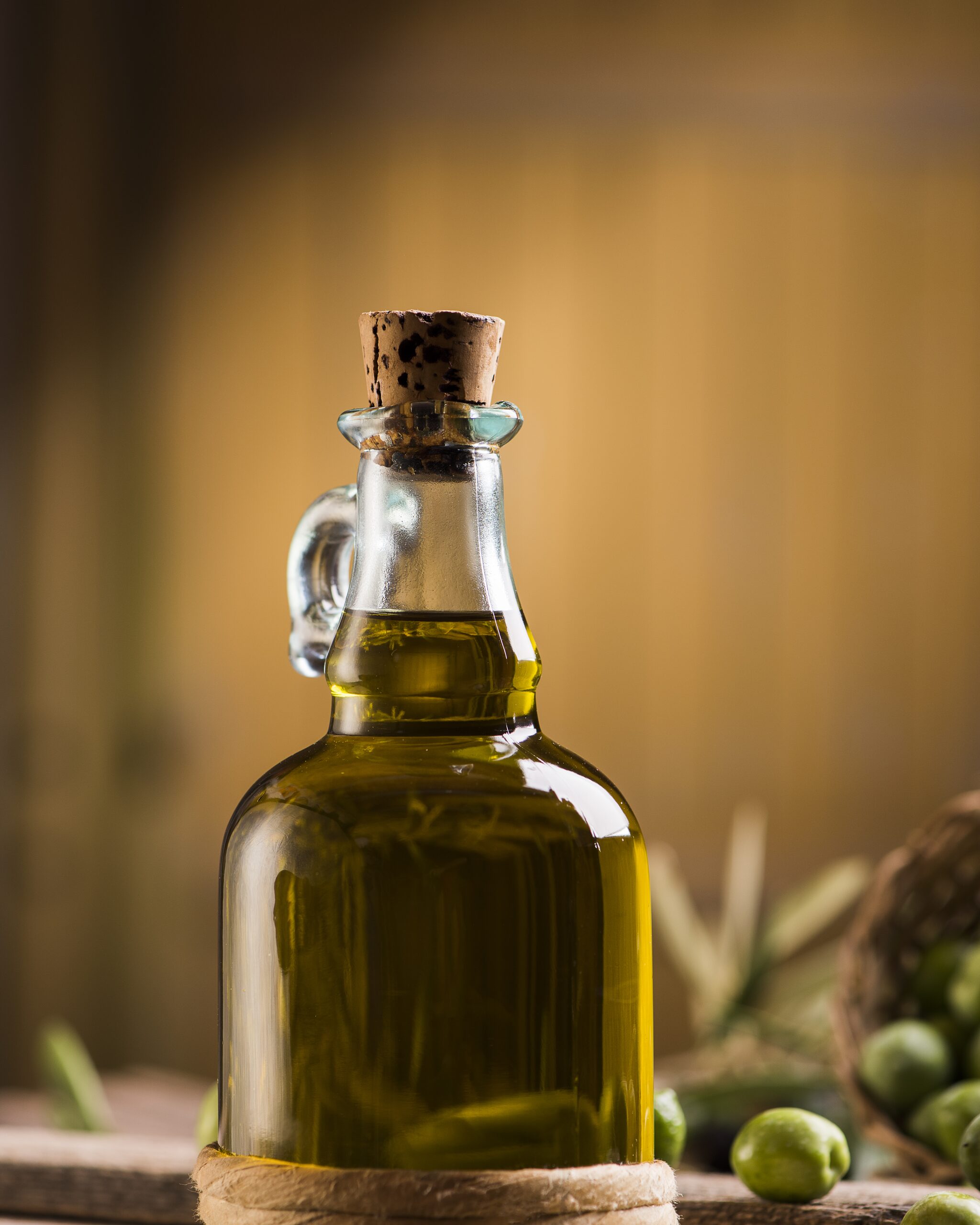 A corked glass bottle of olive oil standing on a wooden surface, with green olives and a basket out of focus behind it.